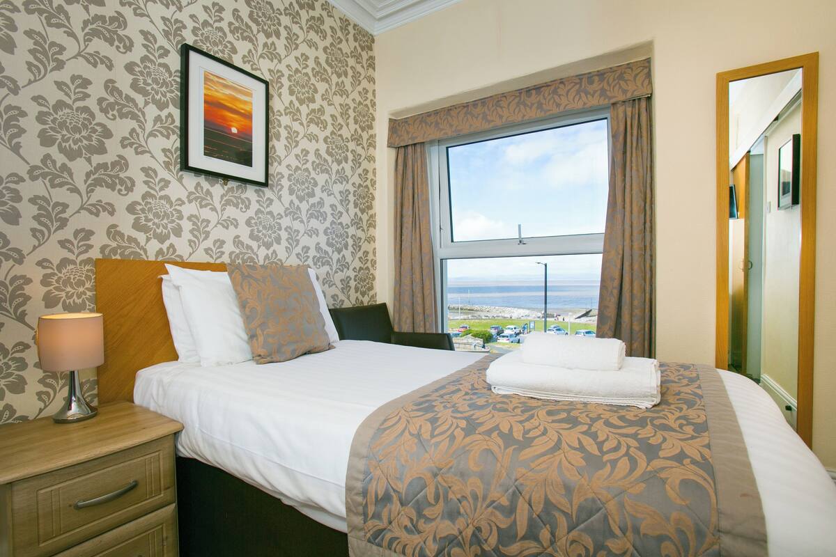 A hotel room with a bed, a nightstand, a lamp, a framed picture on the wall, and a window with a view of the sea.