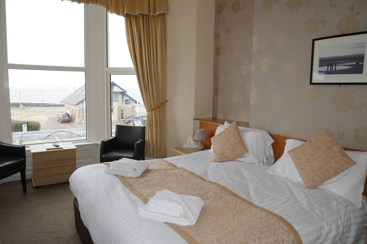 A hotel room with a large bed, a desk, a chair, and a window with a view of the beach and cars.