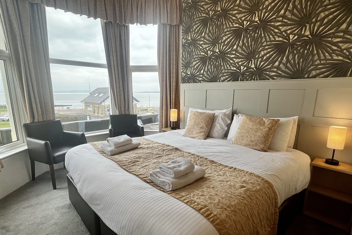 A hotel room with a large bed, a desk, and a chair. There is a view of the sea through the window.