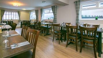 A dining area with multiple tables and chairs, large windows with curtains, and a view of the outside.