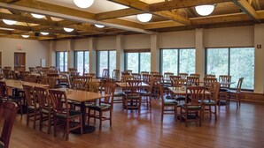 A spacious indoor dining area with wooden tables and chairs, large windows, and a high ceiling with exposed beams.