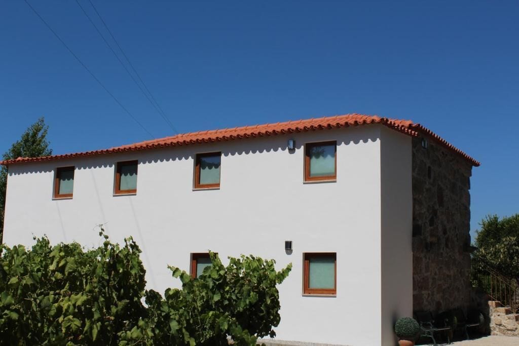 A two-story building with a red-tiled roof and white walls. There are multiple windows with brown frames, and a small tree is visible in front of the building.