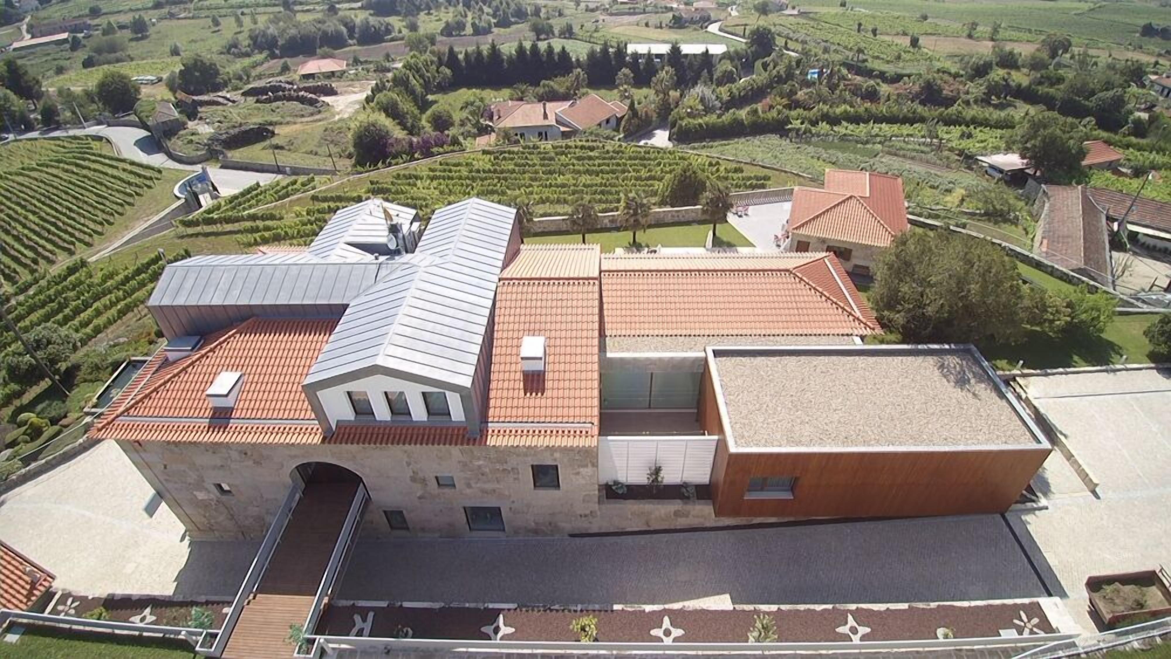Un edificio ampio, su più piani, con un mix di tetti in tegole rosse e lamiera, circondato da giardini curati e vigneti.