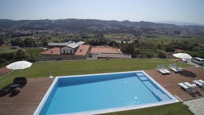 A swimming pool with a wooden deck, surrounded by lounge chairs and umbrellas, with a view of a residential area and hills in the background. - Hotel Rural Quinta das Quintães (Penafiel)