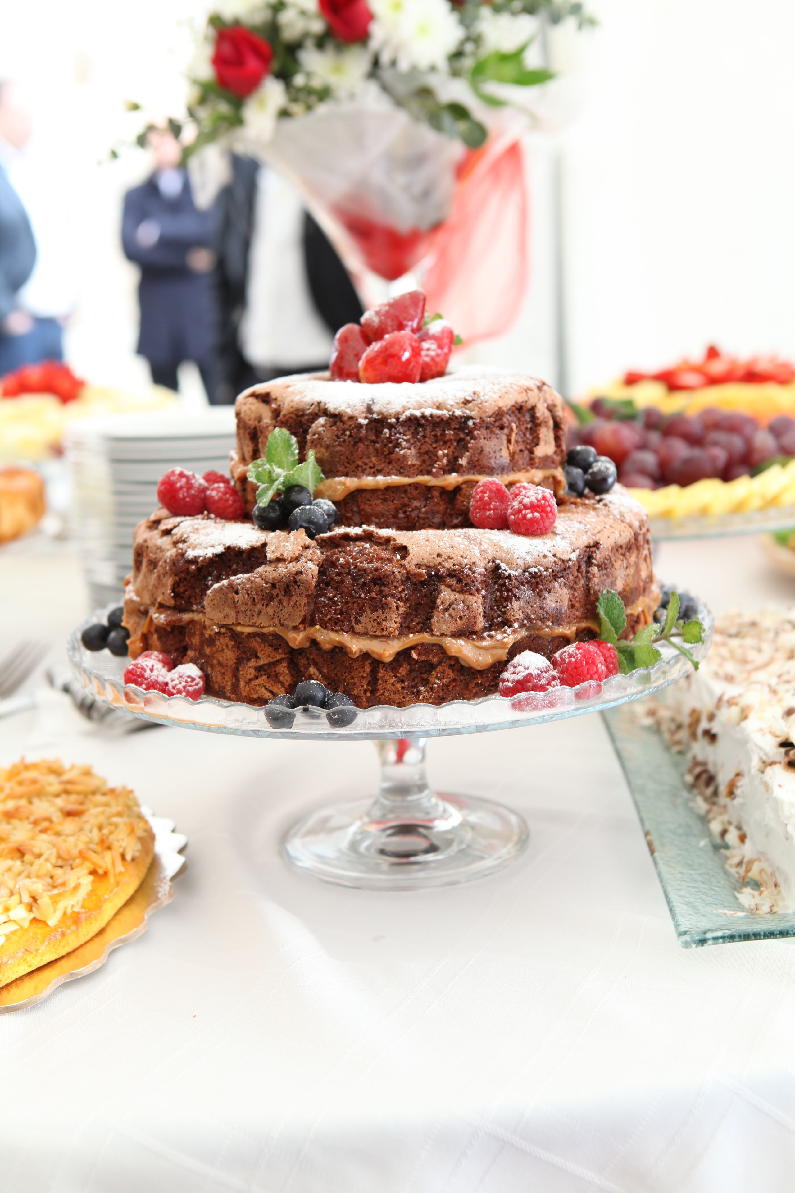 Un pastel de chocolate de varios niveles con adornos de frutas sobre un pedestal de vidrio.