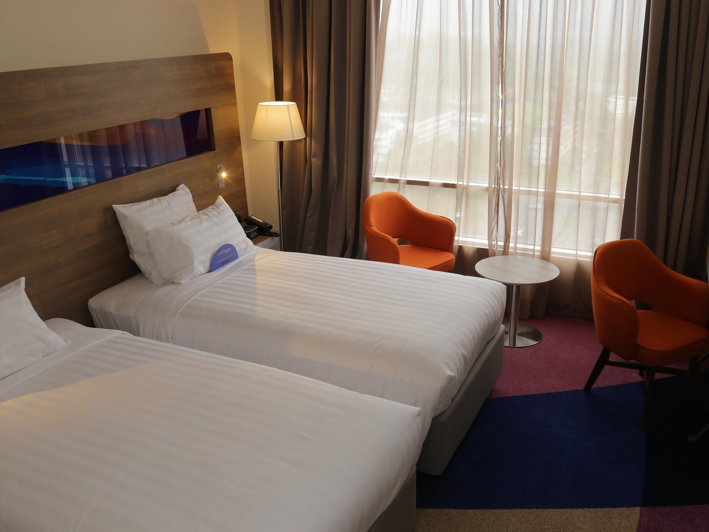 Une chambre d’hôtel avec deux lits, une table de chevet, une lampe, deux fauteuils orange, une petite table ronde et une fenêtre avec des rideaux.