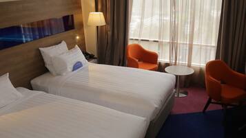 Une chambre d’hôtel avec deux lits, une table de chevet, une lampe, deux fauteuils orange, une petite table ronde et une fenêtre avec des rideaux.