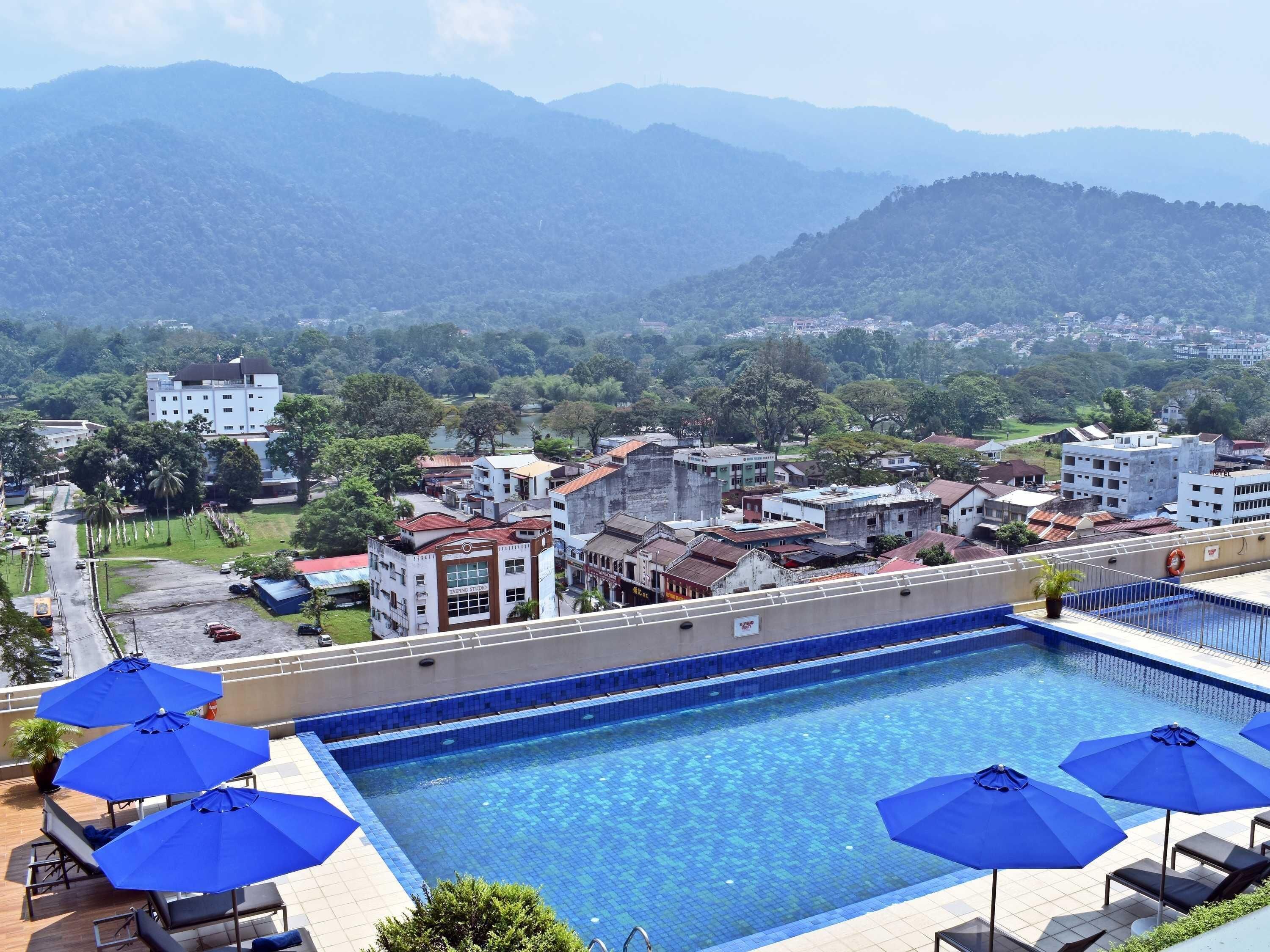 Ein Swimmingpool mit blauen Sonnenschirmen, mit Blick auf eine Stadt und Berge im Hintergrund.