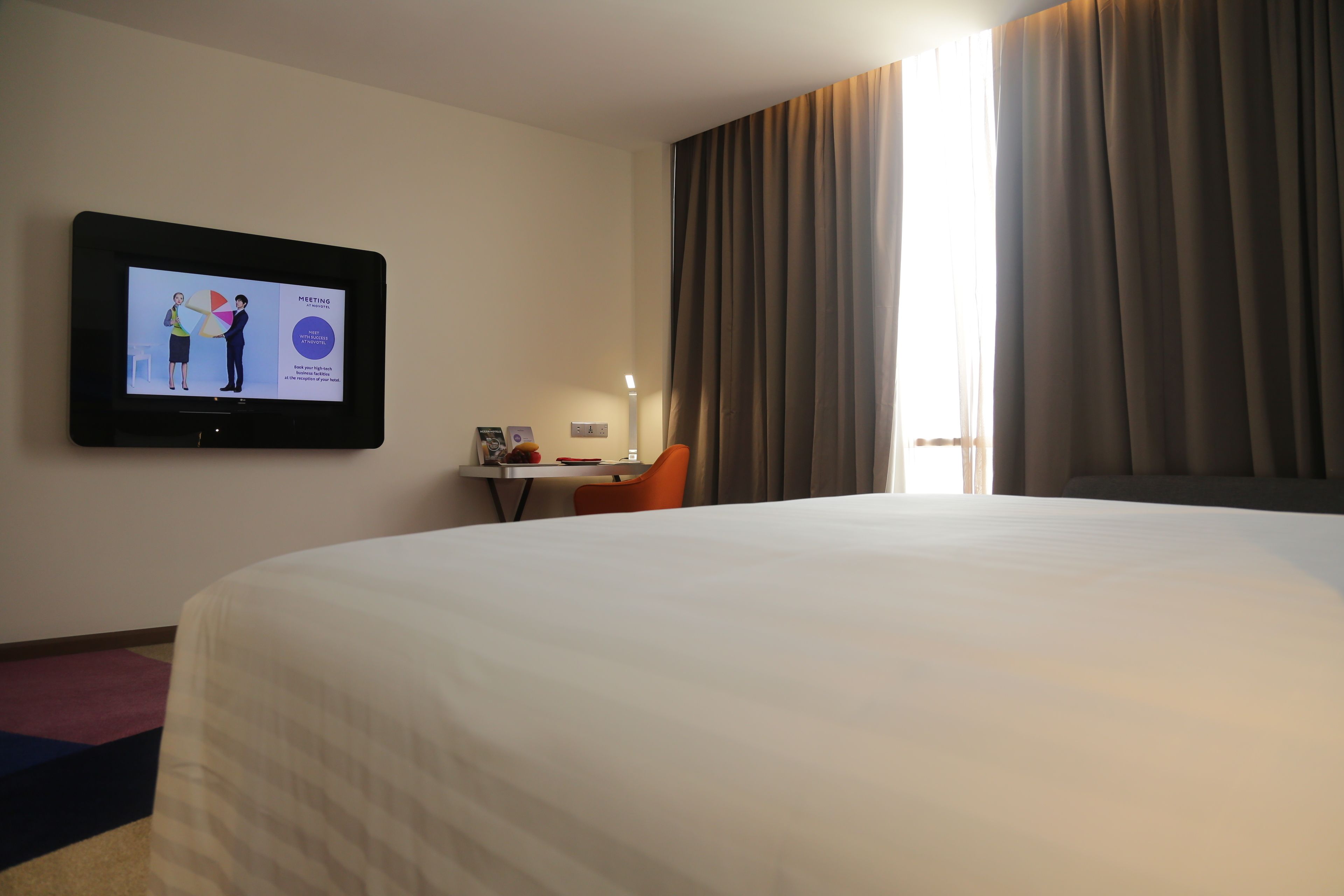 Habitación de hotel con una cama grande, un escritorio con silla, un televisor mostrando una reunión y una ventana con cortinas.