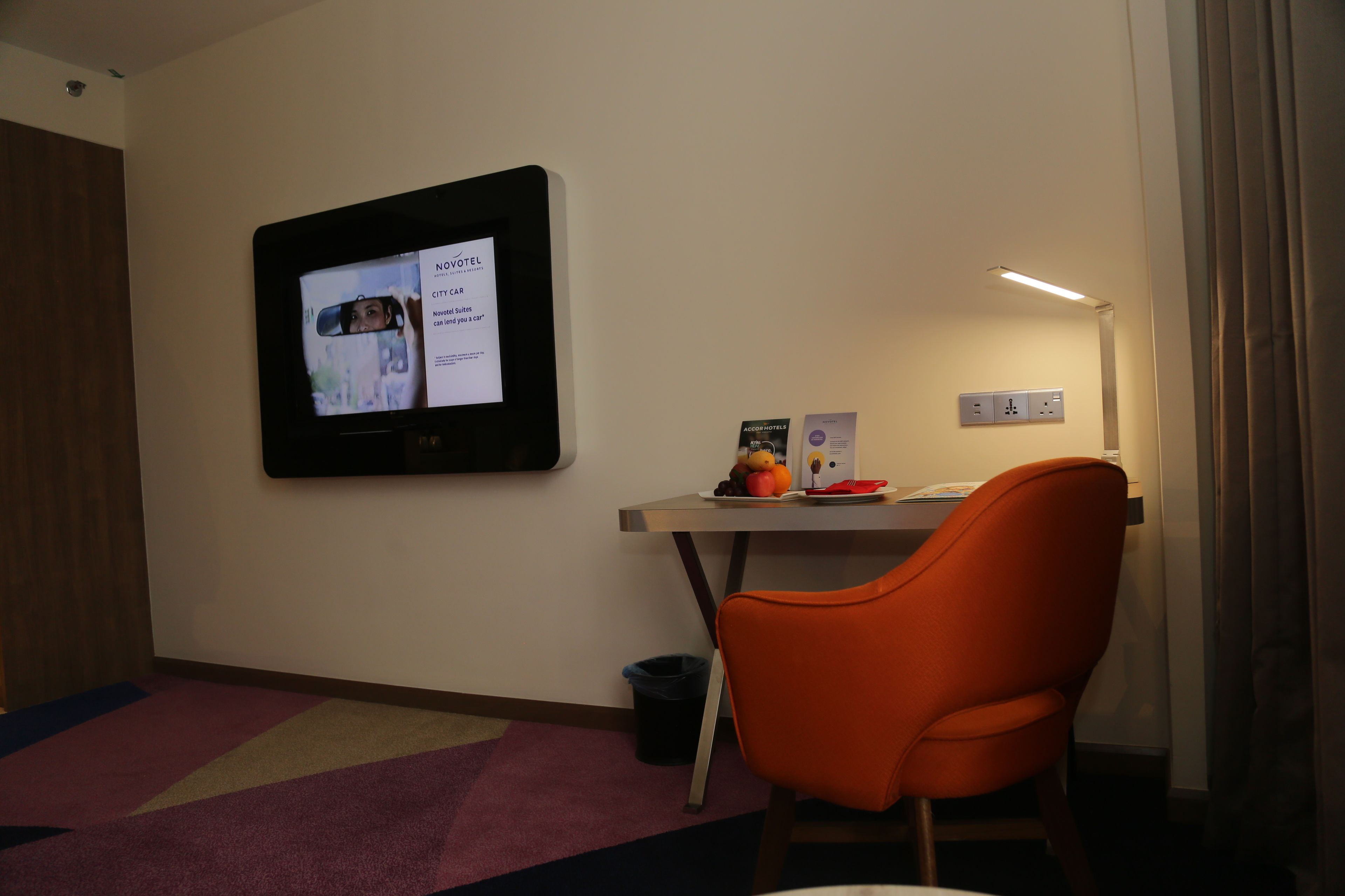 A hotel room with a flat-screen TV mounted on the wall, a small table with a chair, a trash bin, and a lamp.