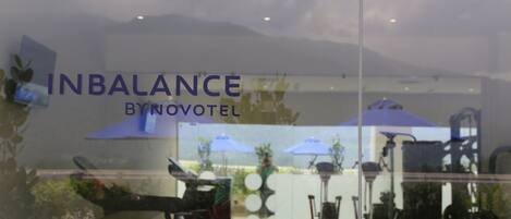 A glass window with the text "INBALANCE BYNOVOTEL" and reflections of outdoor furniture and umbrellas.