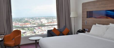 A hotel room with a large bed, a desk, an orange chair, a small round table, and a view of the city through large windows.