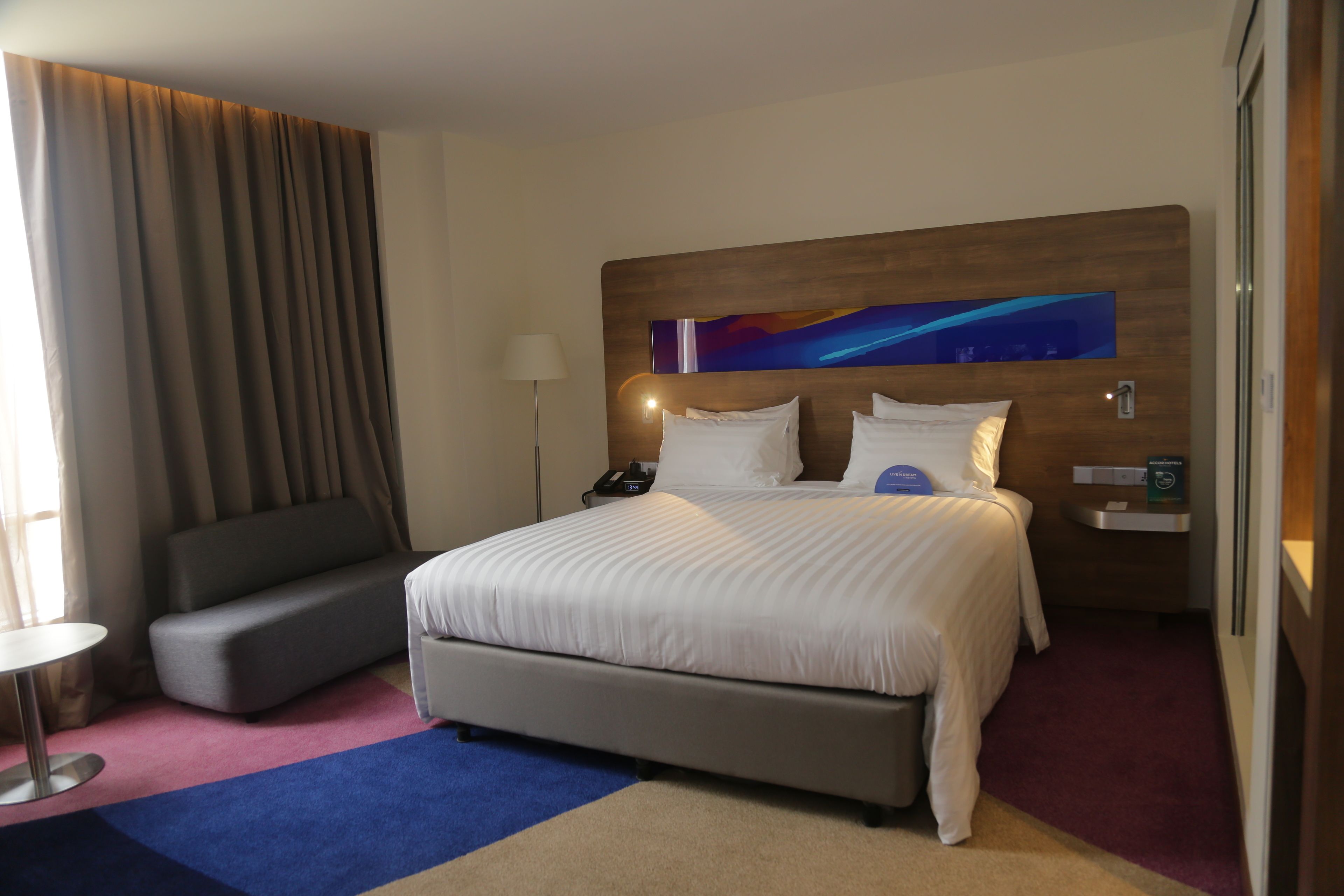 Una habitación de hotel con una cama grande, un sofá gris, una mesita y una mesita de noche con lámpara y teléfono.