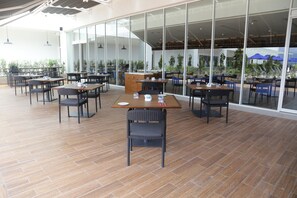 Un amplio espacio exterior para comer con varias mesas y sillas, grandes ventanales y plantas en macetas.