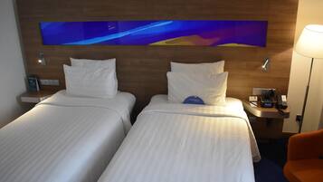 A hotel room with two beds, a nightstand with a phone and a lamp, and a wall-mounted TV.