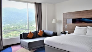 Ein Hotelzimmer mit einem großen Bett, einer Couch mit Kissen, einem Nachttisch und Blick auf eine gebirgige Landschaft durch ein großes Fenster.