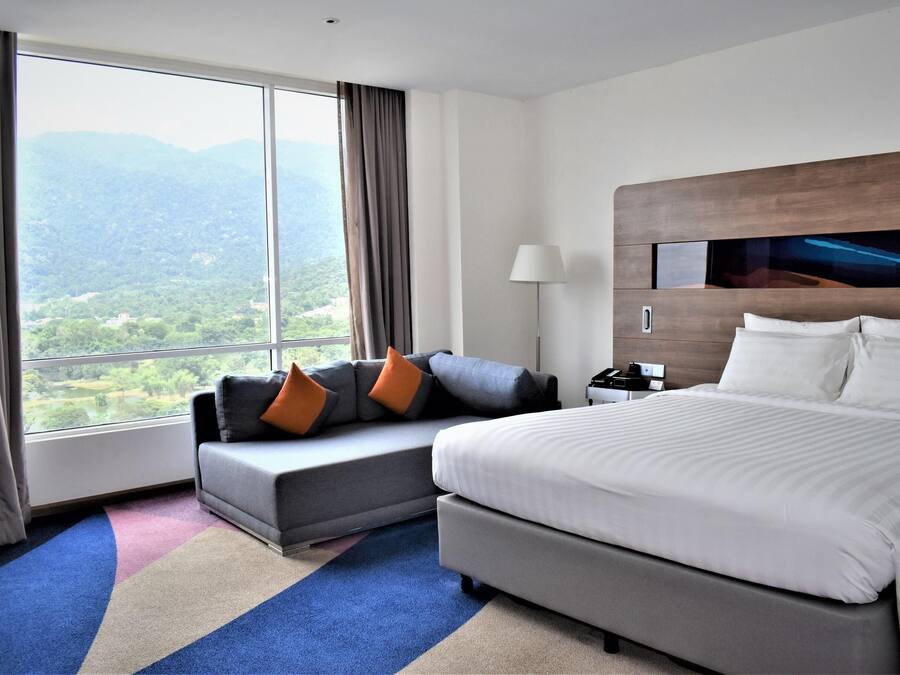 A hotel room with a large bed, a sofa with cushions, a nightstand, and a view of a mountainous landscape through a large window.