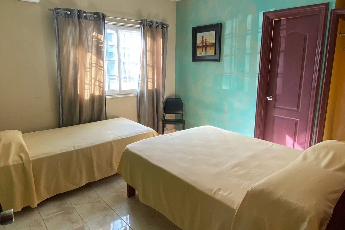 A hotel room with two beds, a window with curtains, a picture on the wall, and a door.