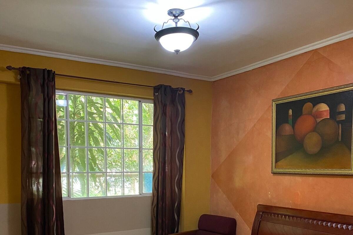 A hotel room with a bed, a chair, a ceiling fan, a window with curtains, and a painting on the wall.