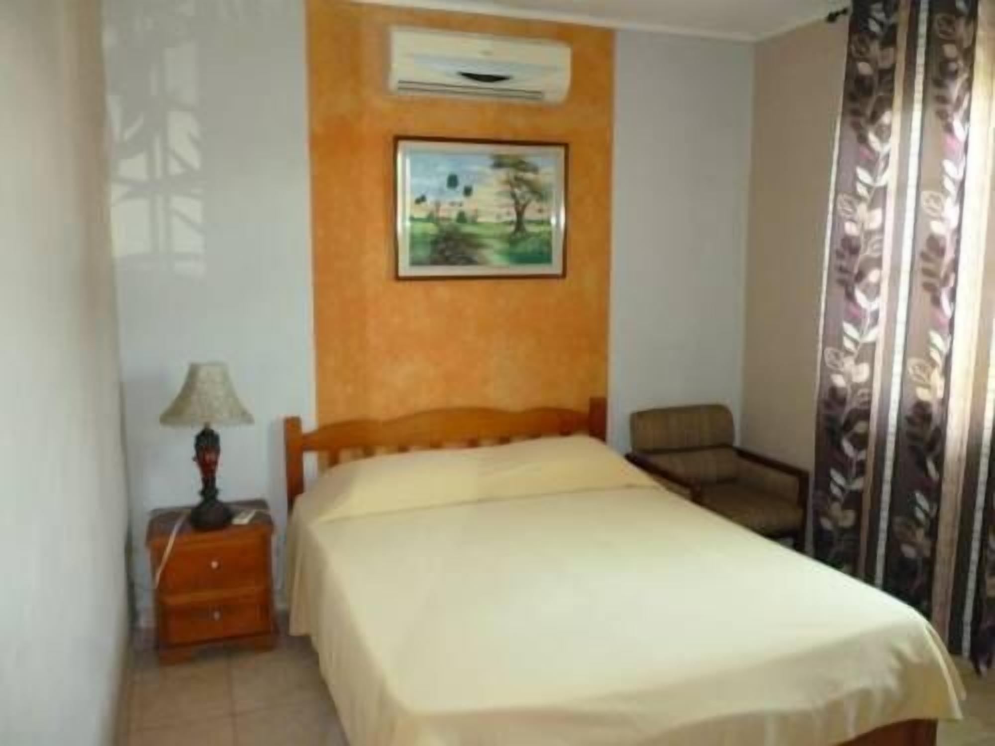 Un dormitorio con cama, mesita de noche, silla, un cuadro en la pared y ventana con cortinas.