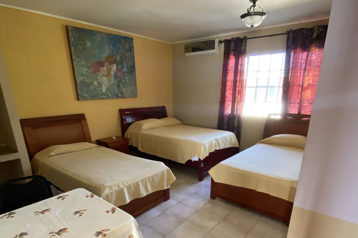 A hotel room with three single beds, a painting on the wall, a window with curtains, and a ceiling fan.