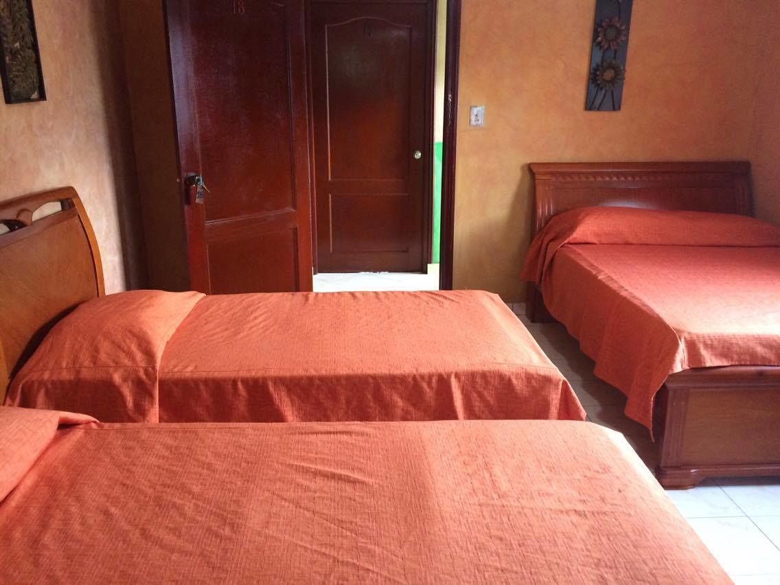 Une chambre avec deux lits, chacun doté d’une tête de lit en bois et recouvert d’une couverture rouge. On aperçoit une porte en bois au fond.