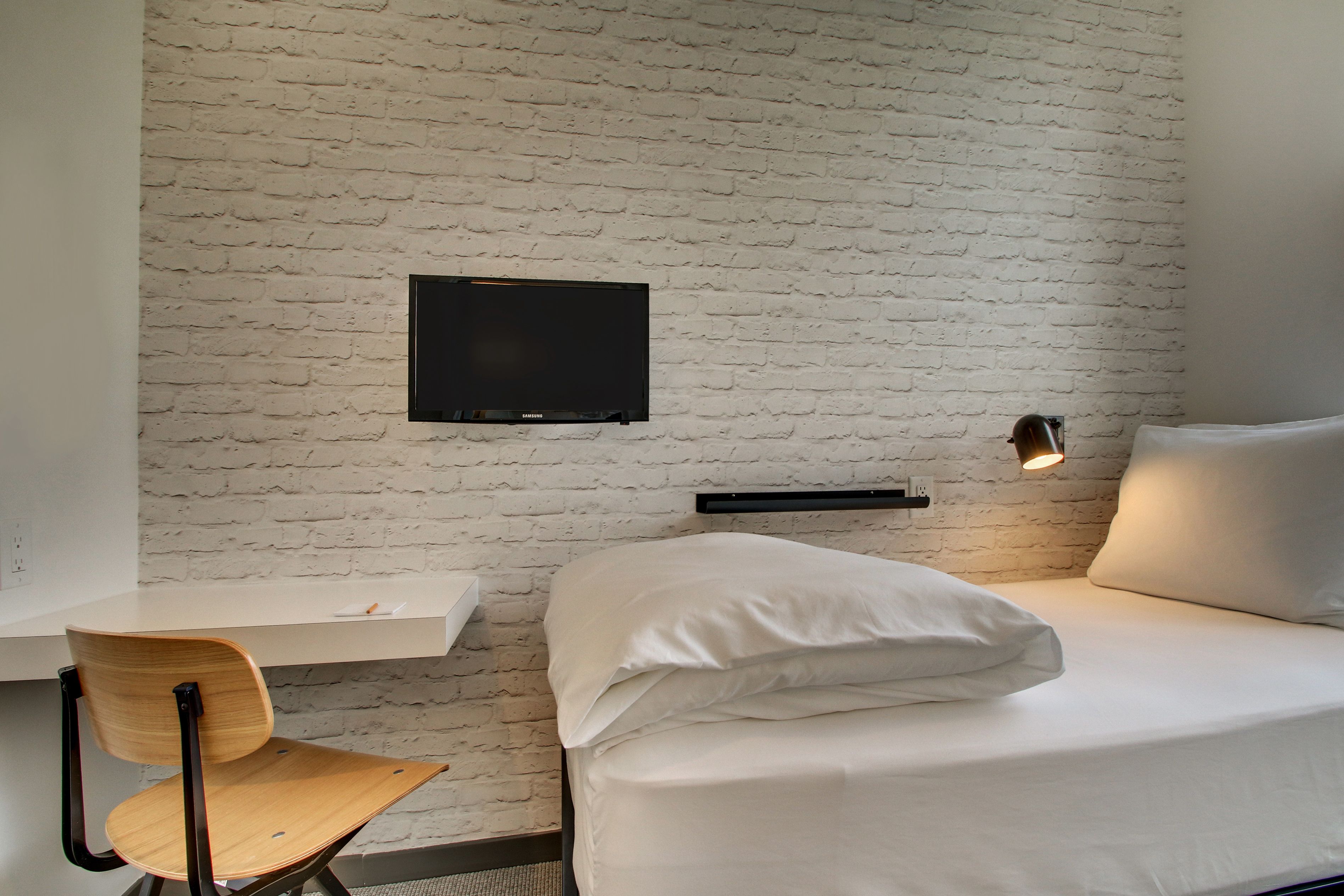A room with a white brick wall, a bed with white bedding, a wooden chair, a flat-screen TV mounted on the wall, and a bedside table with a lamp.