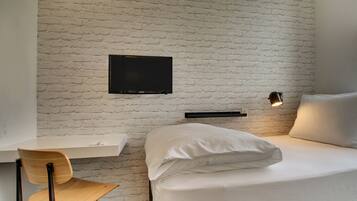 A room with a white brick wall, a bed with white bedding, a wooden chair, a flat-screen TV mounted on the wall, and a bedside table with a lamp.