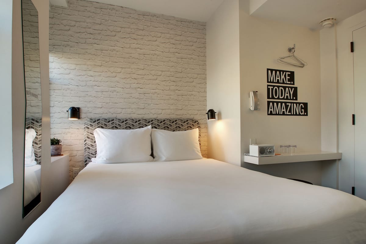A modern hotel room with a brick-patterned wall, a bed with white linens, a bedside table with a lamp, and a motivational quote on the wall.