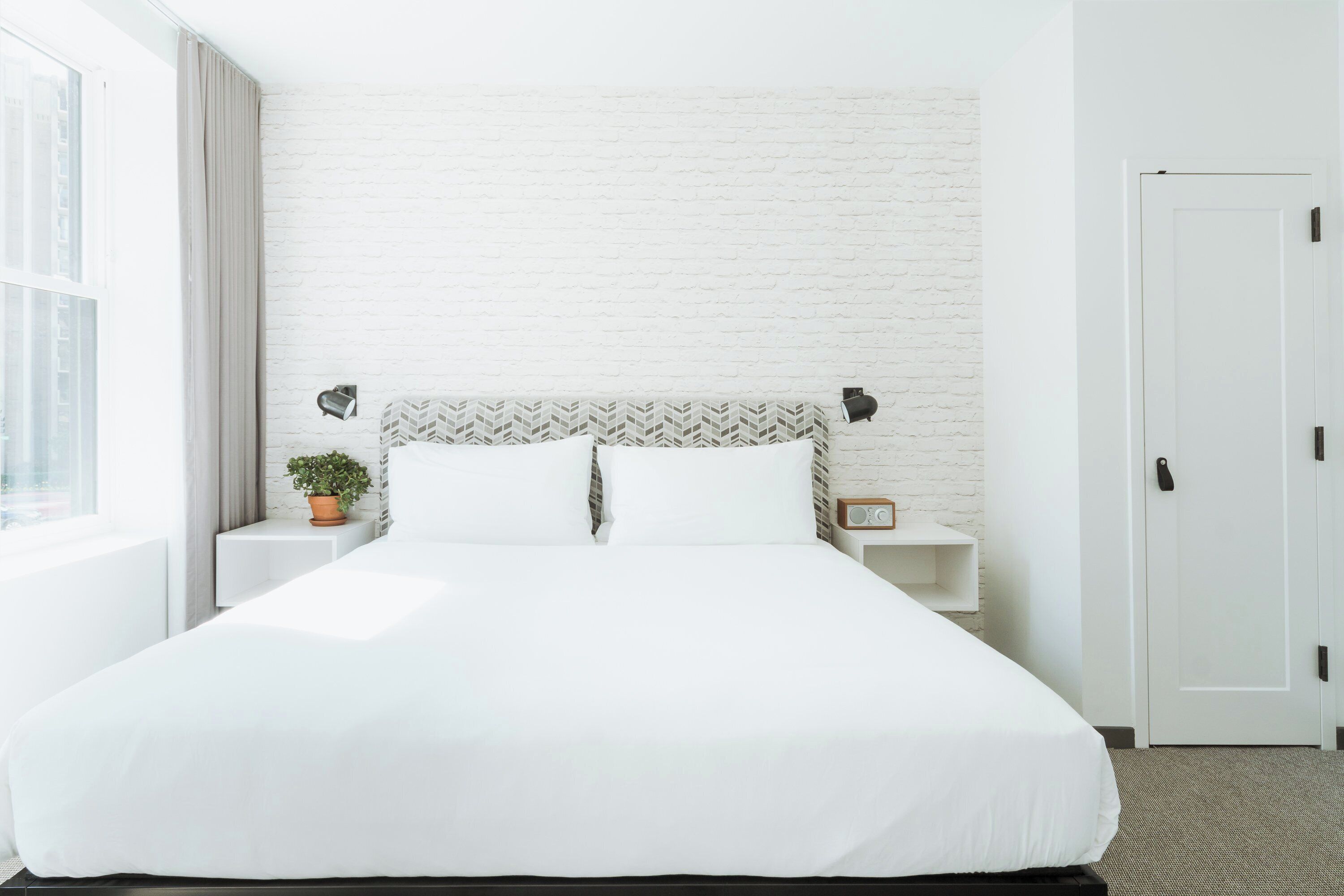 A neatly made bed with white bedding, a white brick wall, a potted plant, and a bedside table with a radio.