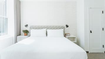 A neatly made bed with white bedding, a white brick wall, a potted plant, and a bedside table with a radio.
