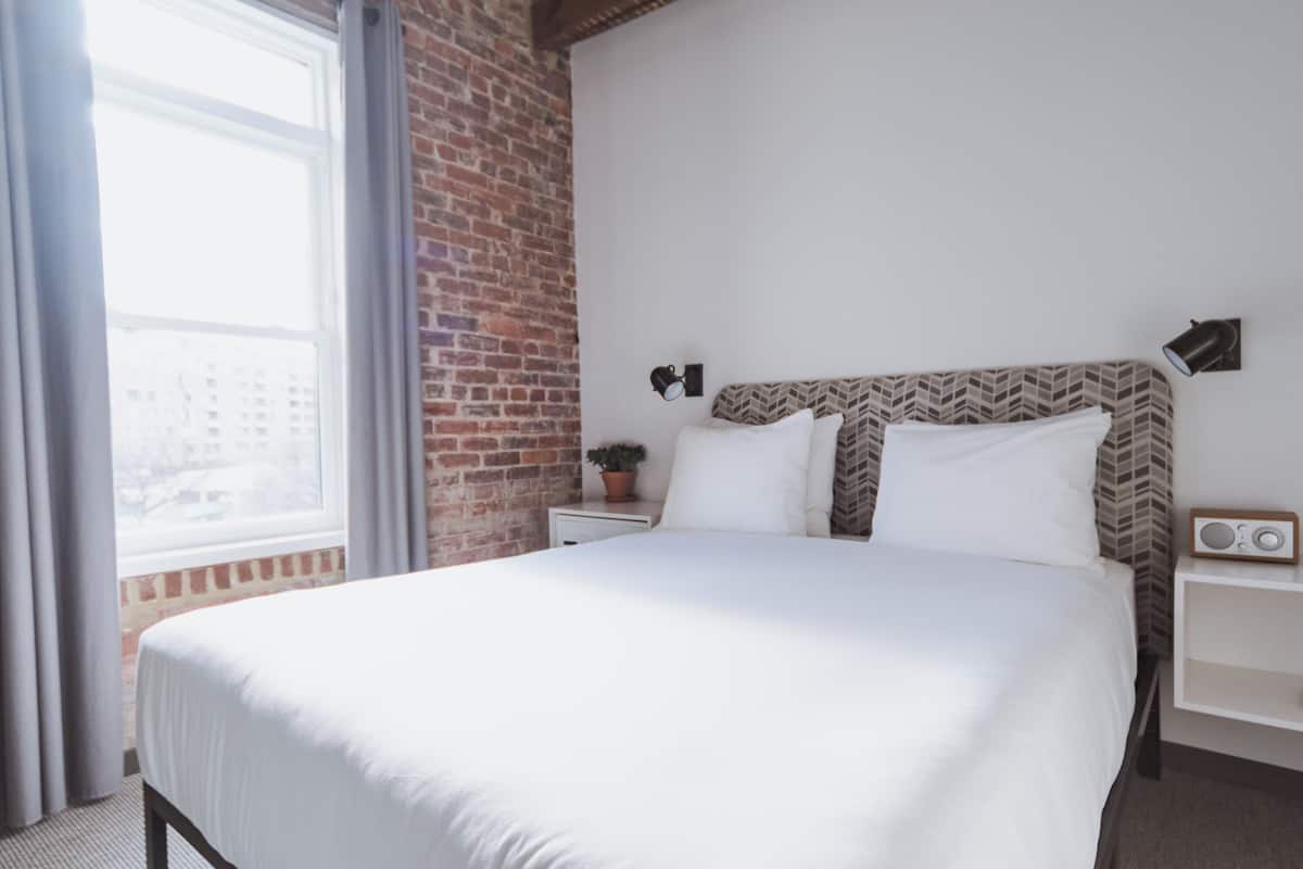 A neatly made bed in a hotel room with a brick wall, a small bedside table with a radio, and a window with curtains.