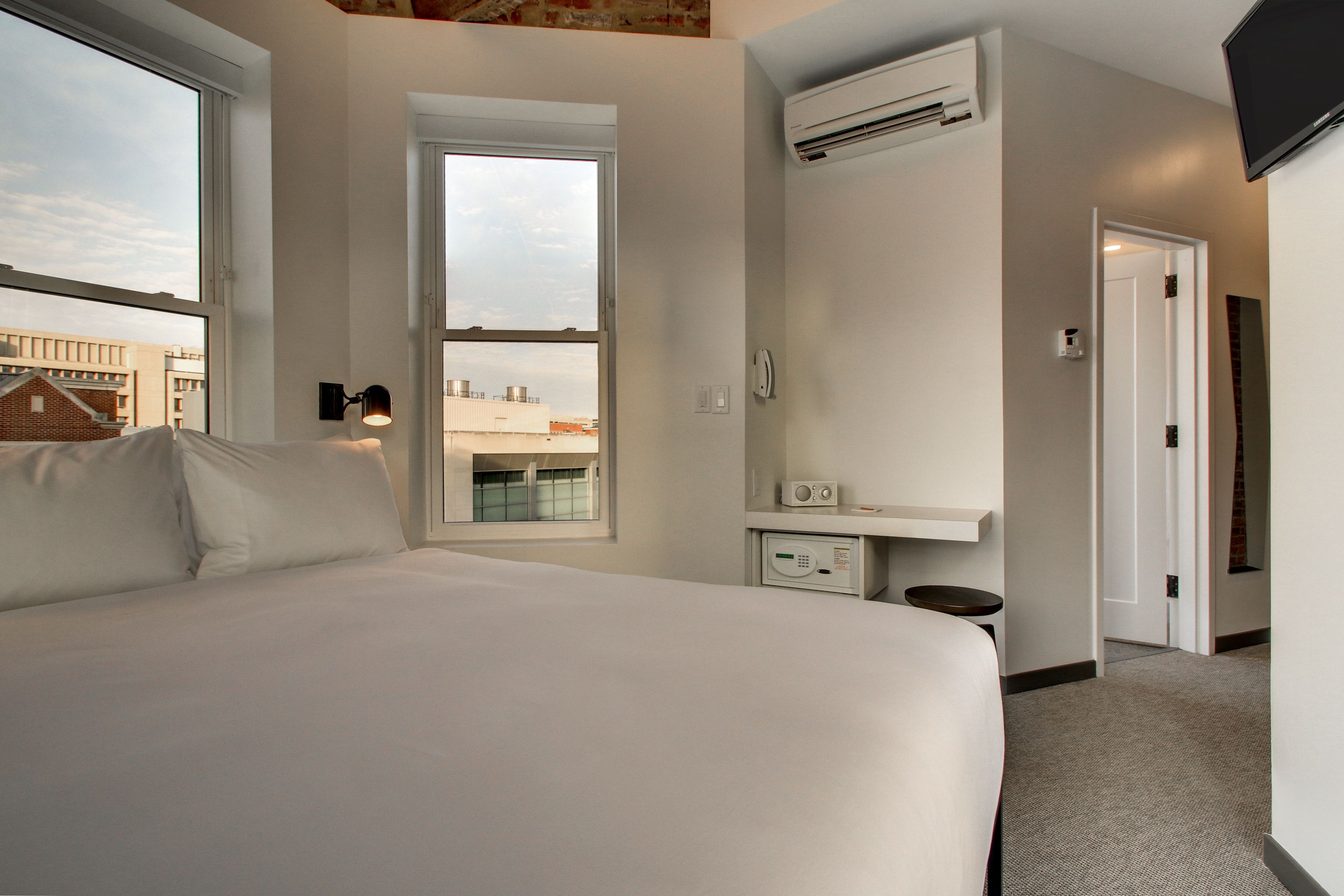 A hotel room with a large bed, a window, an air conditioning unit, a bedside table, and a small table with a radio.