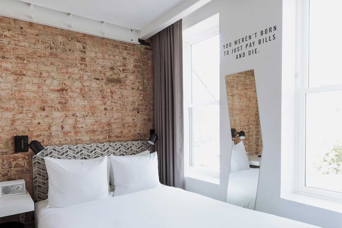 A hotel room with a brick wall, a bed with white bedding, a desk with a radio, and a window with curtains.
