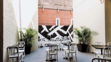 Un espace extérieur aménagé avec des tables et des chaises, des plantes en pot et un mur de briques orné d’une fresque géométrique.