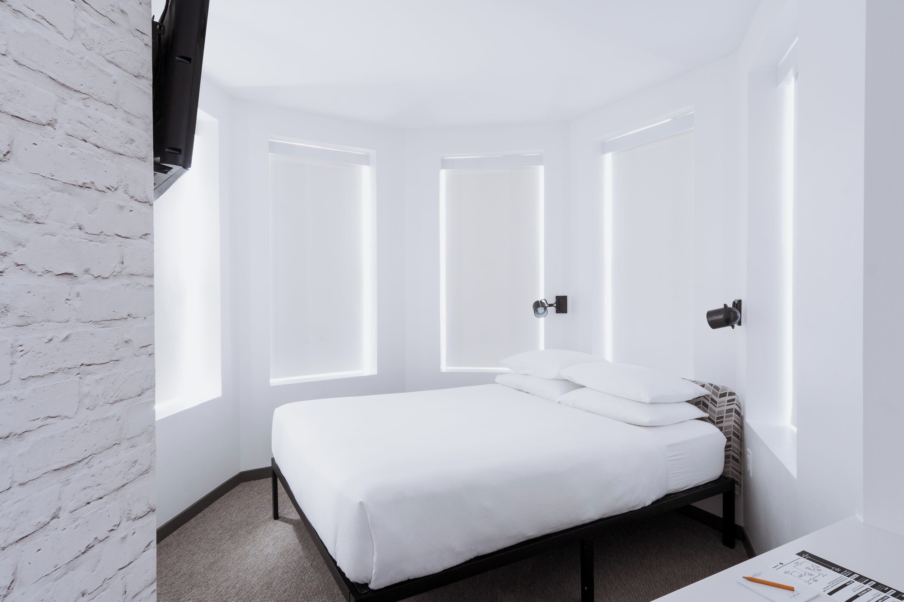 Una habitación de hotel moderna con una pared de ladrillo blanco, una cama bien hecha y ventanas grandes con iluminación integrada.