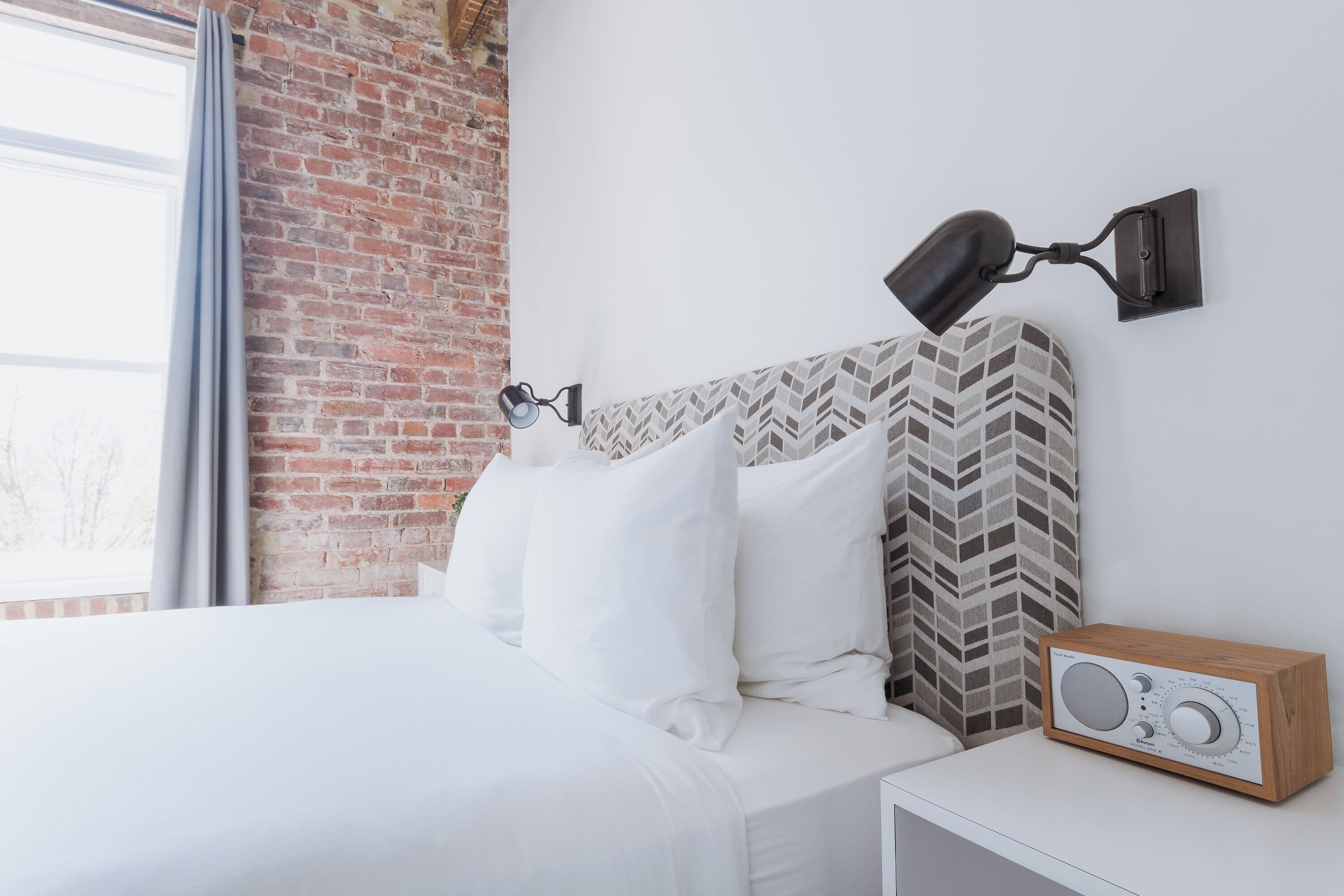Habitación de hotel con pared de ladrillo, cama con ropa blanca, cabecera estampada, mesita de noche con radio y ventana con cortinas.
