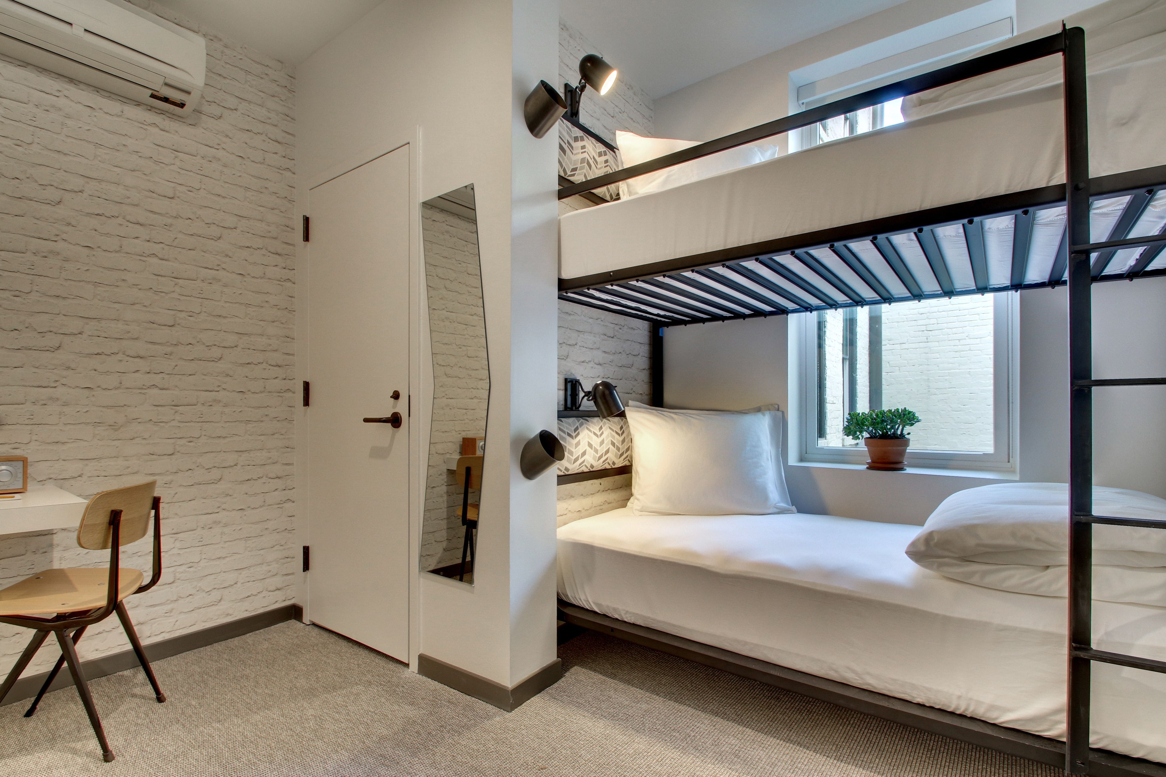 Un lit superposé avec un matelas blanc, un bureau avec une chaise et une plante en pot près de la fenêtre.