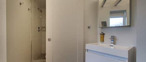 Une salle de bain moderne avec une douche à l’italienne en verre, un lavabo blanc avec un miroir au-dessus, et une porte donnant sur une autre pièce.