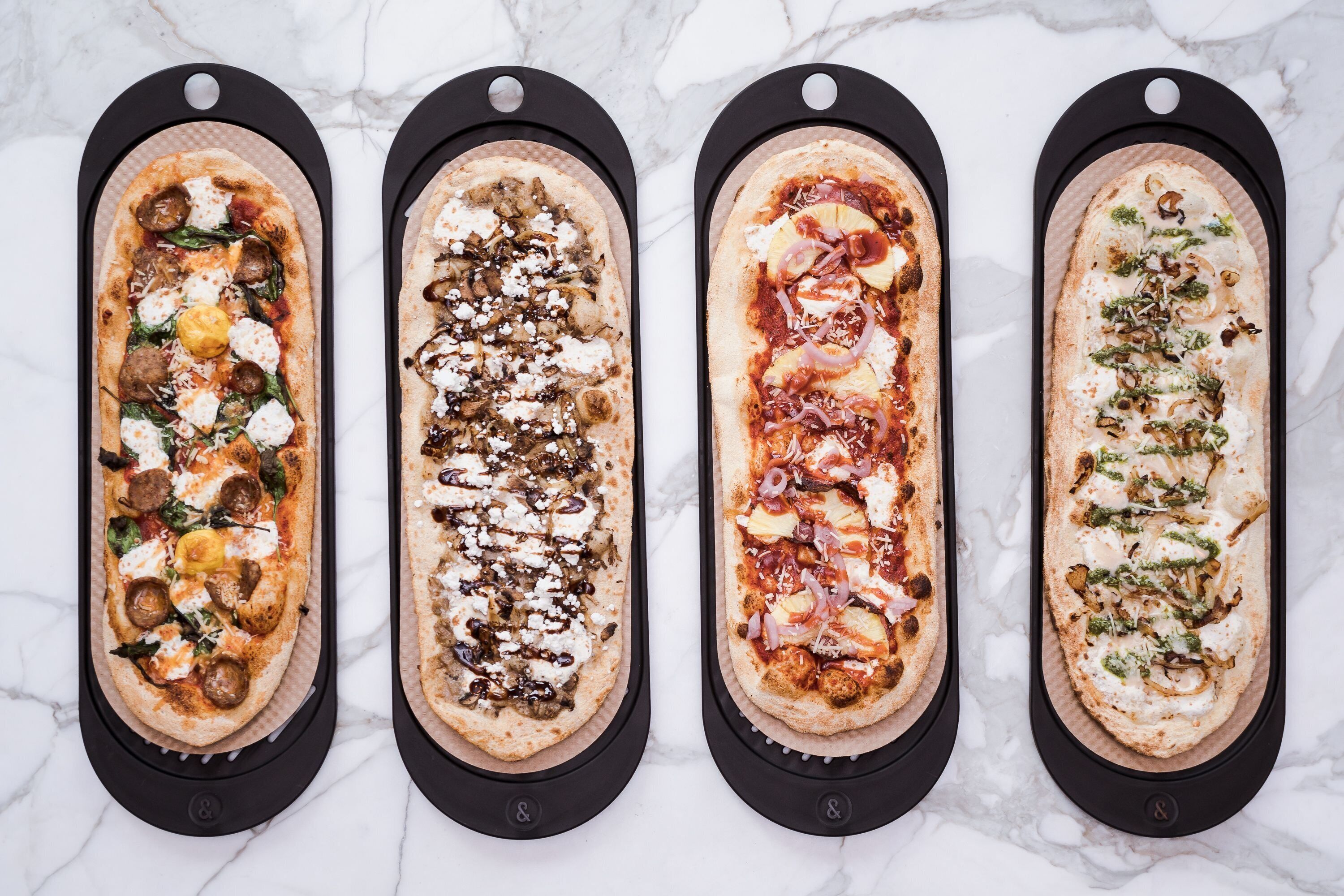 Cuatro pizzas sobre bandejas negras con distintos ingredientes, dispuestas sobre una superficie de mármol.
