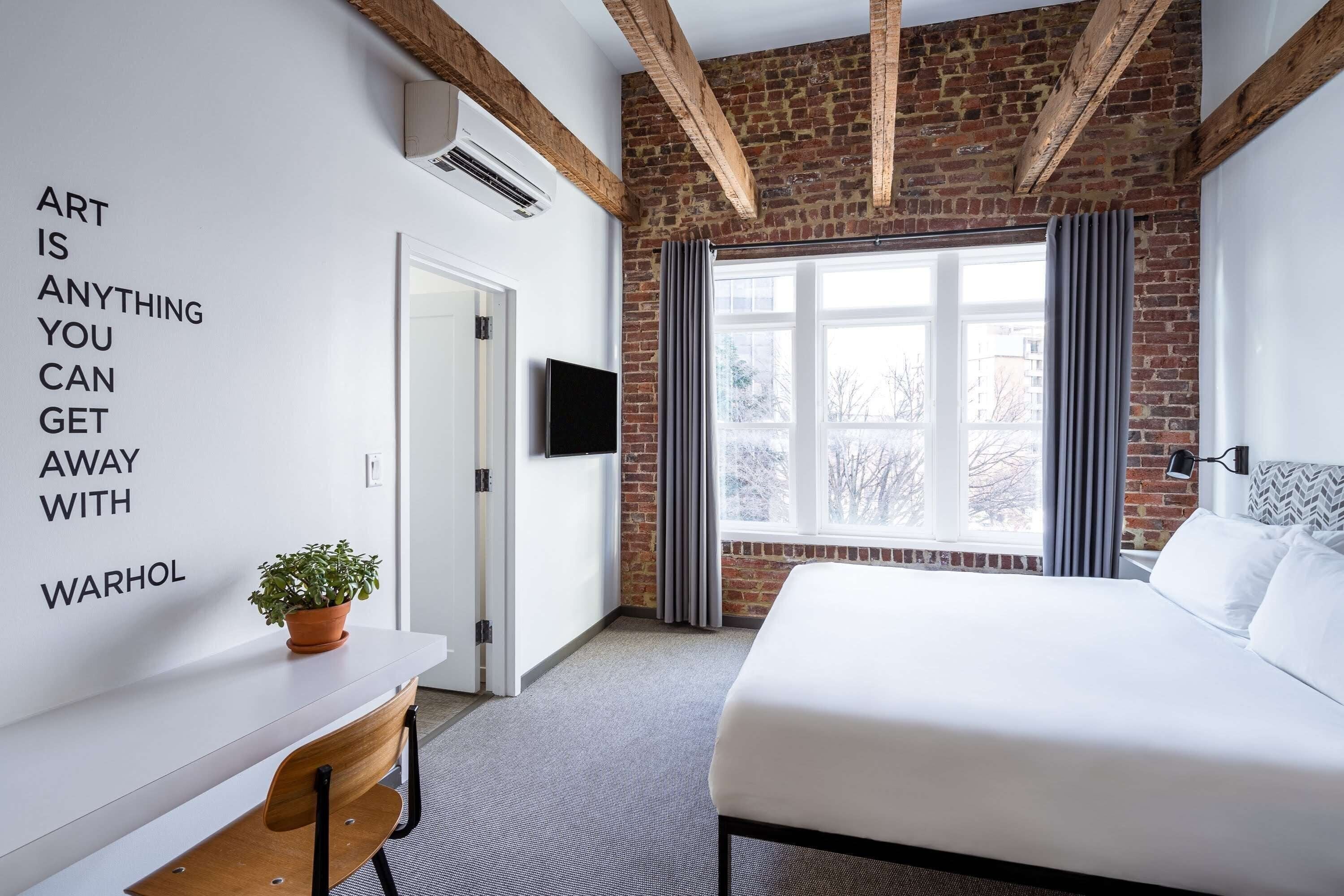 Ein modernes Hotelzimmer mit freiliegenden Backsteinwänden, einem großen Fenster mit Vorhängen, einem Bett, einem Schreibtisch mit Pflanze und einem motivierenden Zitat an der Wand.