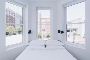 Une chambre à coucher avec un lit placé au centre, entre deux fenêtres, offrant une vue sur des immeubles et une rue.