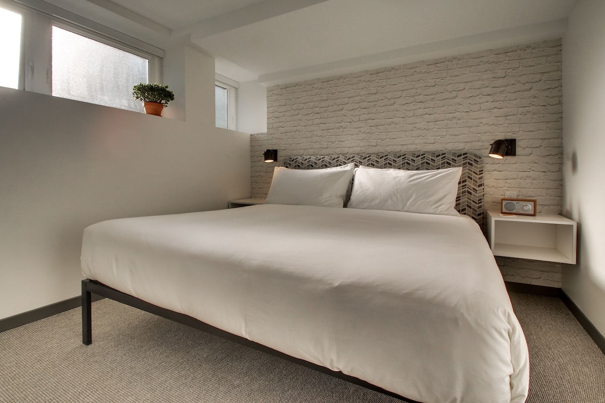 A modern bedroom with a large bed, a brick-patterned headboard, bedside lamps, and a small plant.