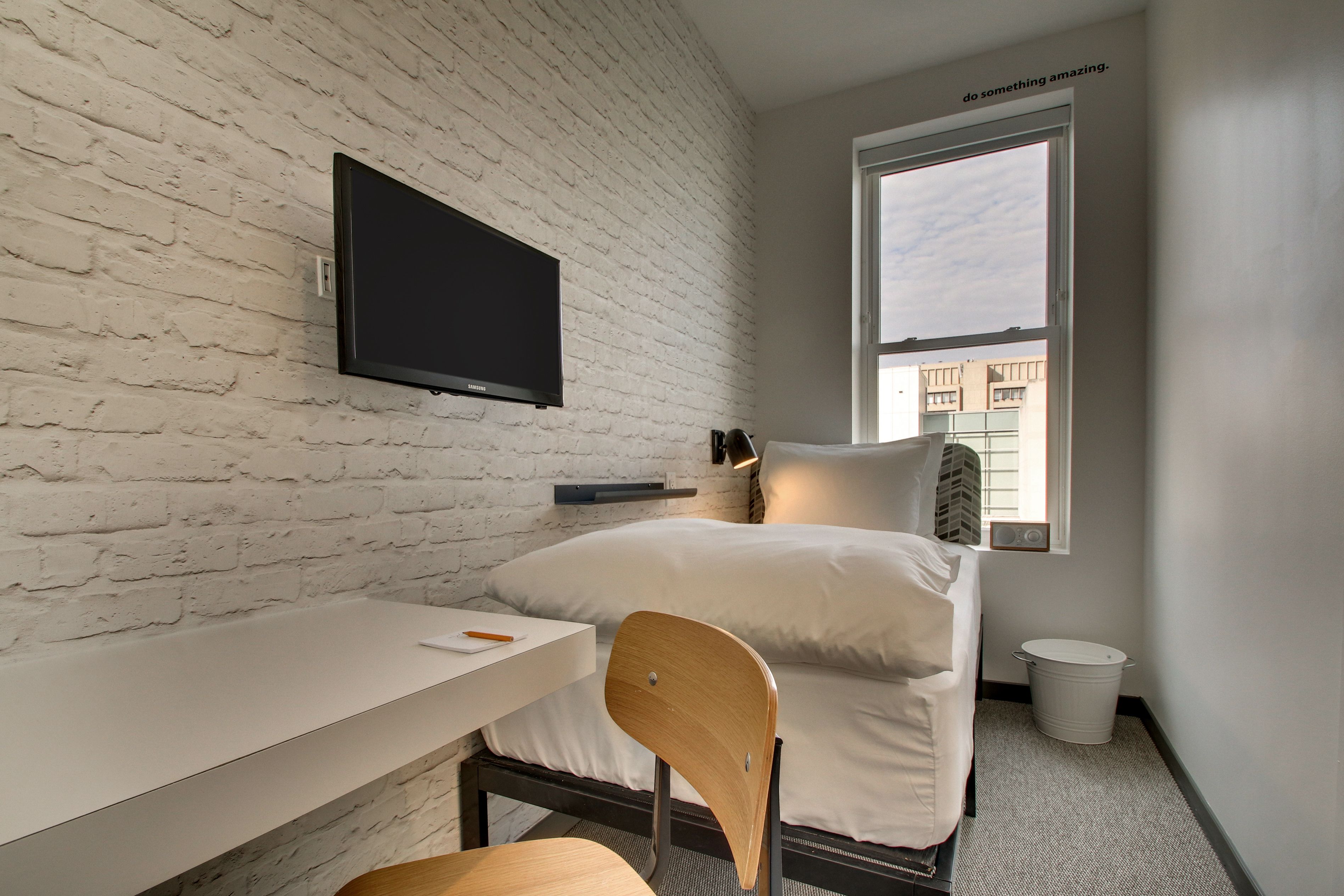 A modern hotel room with a white brick wall, a flat-screen TV, a bed with white bedding, a small table with a chair, and a window with a view of buildings.