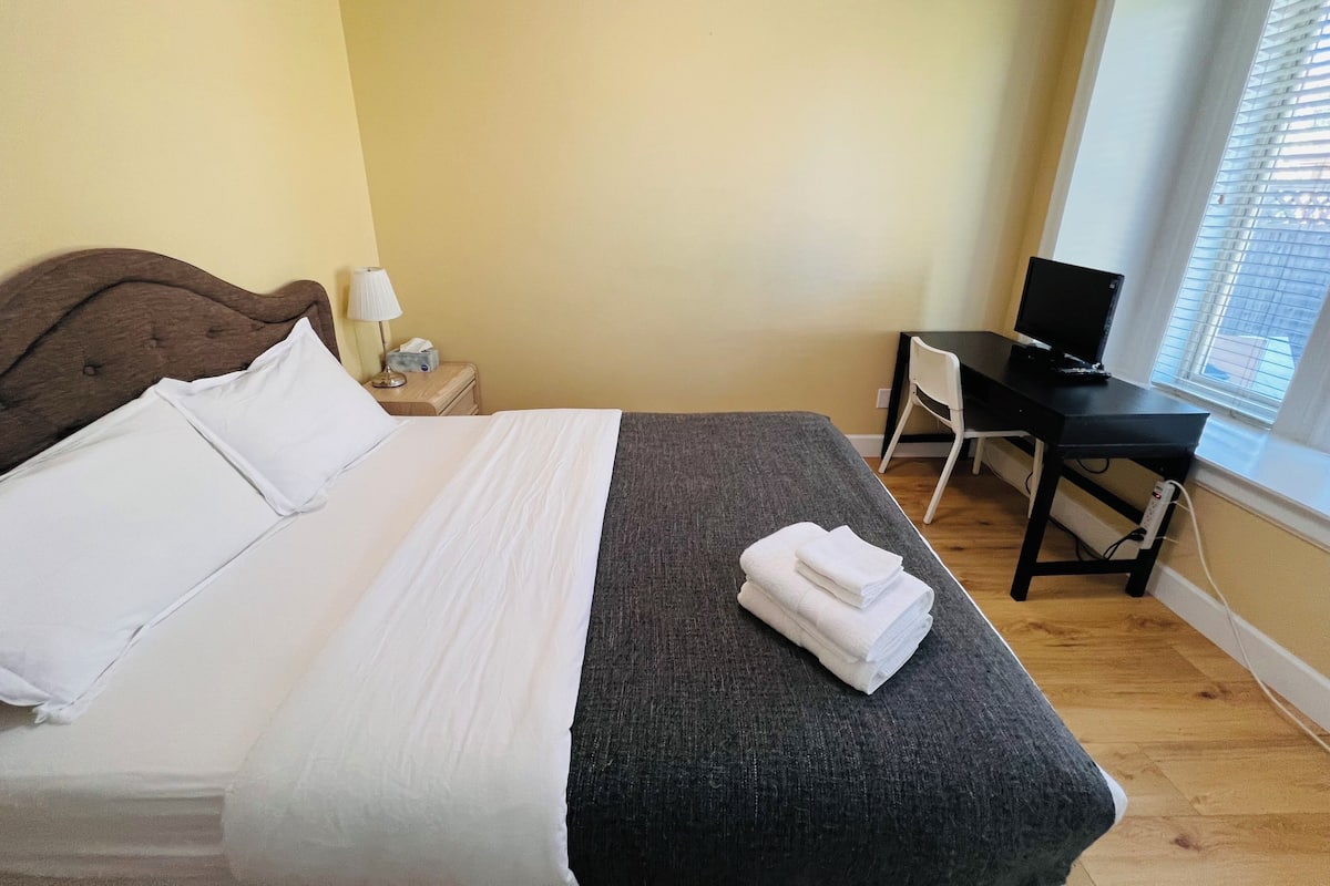 A neatly made bed with white linens, a grey blanket, and a stack of white towels on the foot of the bed. There is a wooden nightstand with a lamp and a small table beside the bed. A black desk with a computer monitor is positioned near a window with blinds.