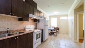 Una cocina con armarios de madera oscura, un horno blanco y un refrigerador. Se ve un comedor con mesa y sillas al fondo.