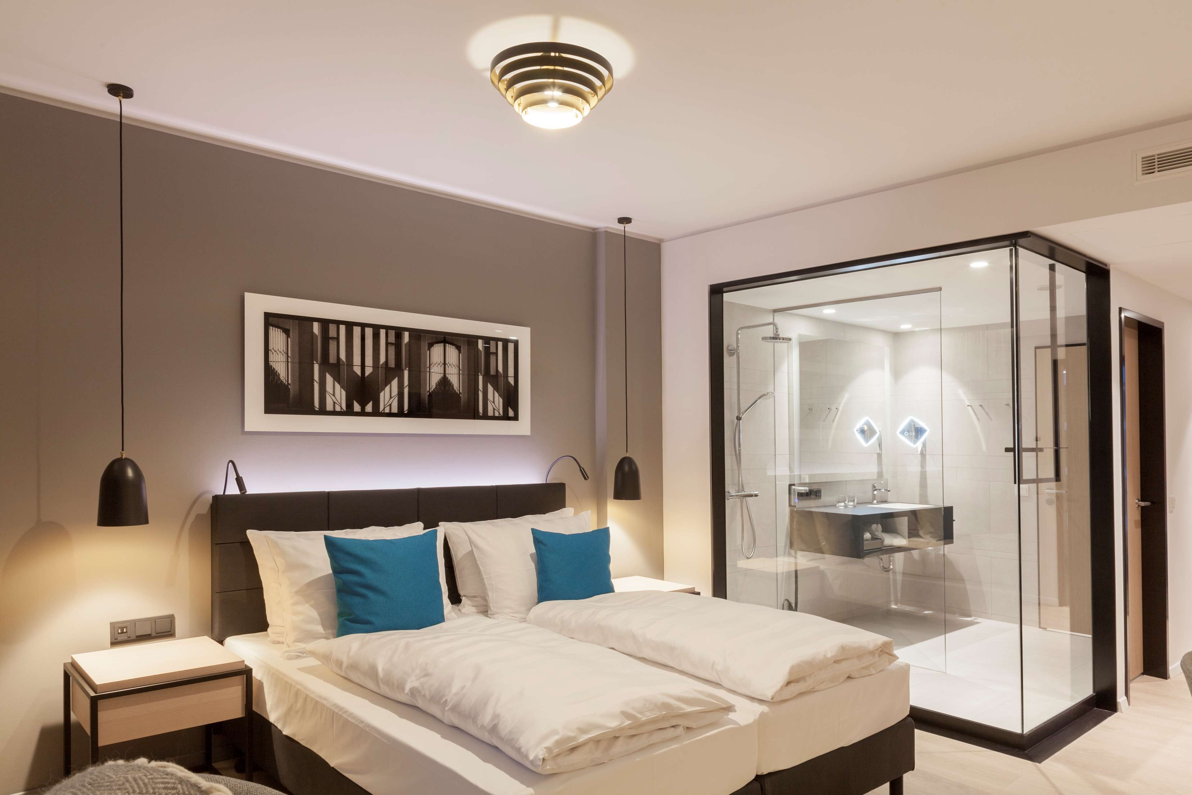 A modern bedroom with a large bed, bedside tables, a nightstand, a framed artwork, and a glass-enclosed shower area.