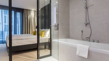 Un dormitorio moderno con una puerta de cristal que da a un baño. La habitación cuenta con una cama grande, un box de ducha con mampara de cristal y una bañera blanca.