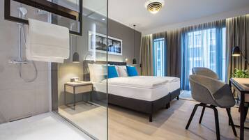 Quarto de hotel moderno com box de vidro, cama com roupa de cama azul e branca, escrivaninha com cadeira e uma janela grande com cortinas.