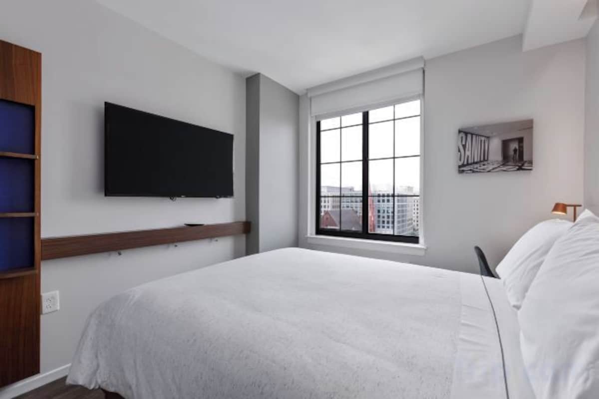 A hotel room with a bed, a television mounted on the wall, a window with a view of buildings, and a small picture frame on the wall.